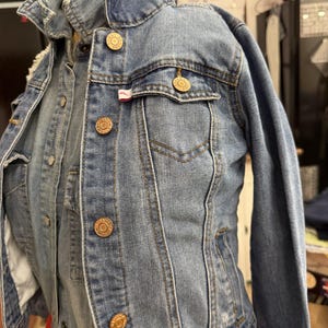 May include: A light blue denim jacket featuring a classic collar, long sleeves, and a button-up front. The jacket includes two chest pockets with buttoned flaps, each adorned with metallic buttons embossed with the text Tommy Hilfiger.