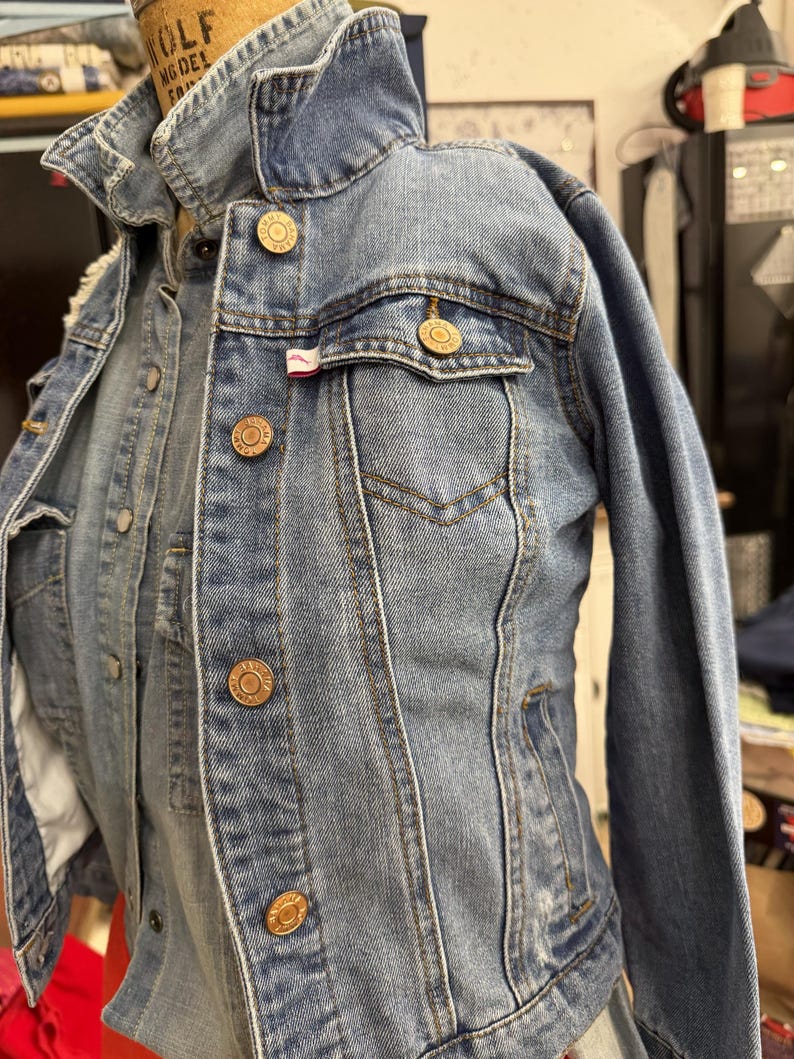 May include: A light blue denim jacket featuring a classic collar, long sleeves, and a button-up front. The jacket includes two chest pockets with buttoned flaps, each adorned with metallic buttons embossed with the text Tommy Hilfiger.