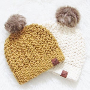 May include: Two crocheted beanies with faux fur pom poms. One beanie is mustard yellow and the other is white. Both beanies have a brown leather tag with the letters "CD" on them.