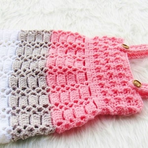 May include: A crocheted baby dress with white, beige, and pink stripes. The dress has two straps with wooden buttons.