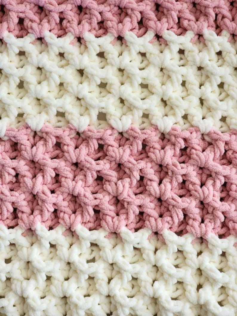 May include: A close-up of a crocheted blanket with alternating stripes of pink and white yarn. The blanket has a textured, open-weave design.