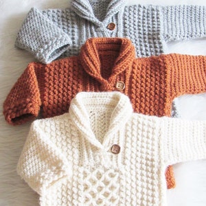 May include: Three hand-knitted baby sweaters in gray, rust, and cream. Each sweater features a shawl collar, long sleeves, and a wooden button closure. The cream sweater has a diamond pattern on the front.