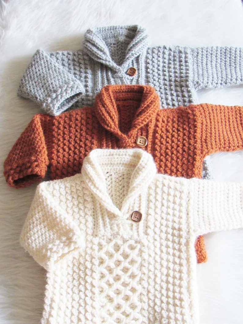 May include: Three hand-knitted baby sweaters in gray, rust, and cream. Each sweater features a shawl collar, long sleeves, and a wooden button closure. The cream sweater has a diamond pattern on the front.