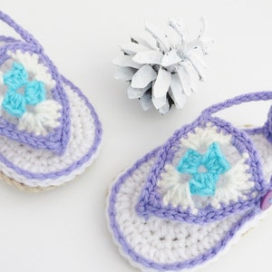 May include: A pair of crocheted baby sandals in white, purple and blue. The sandals feature a flower design and a button fastening.