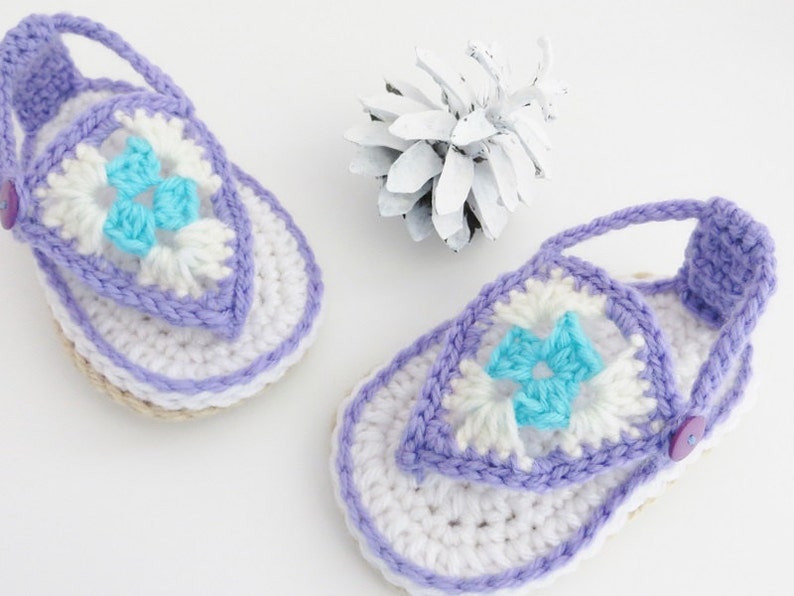 May include: A pair of crocheted baby sandals in white, purple and blue. The sandals feature a flower design and a button fastening.
