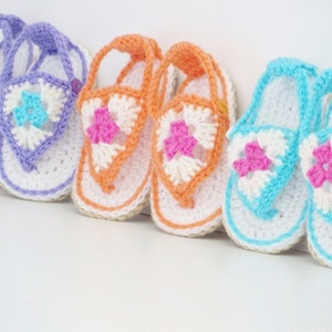 May include: Three pairs of crocheted baby sandals in purple, orange, and blue with white accents. Each sandal features a flower design in the centre.