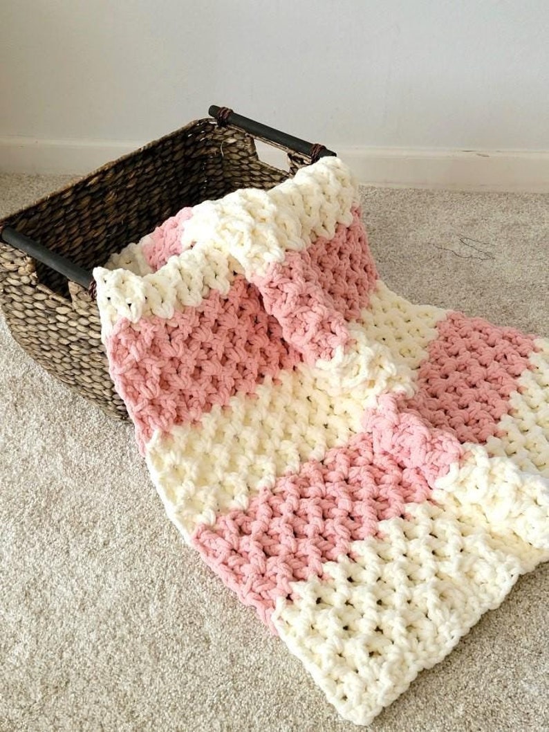 May include: A pink and white striped crocheted blanket with a textured stitch pattern. The blanket is folded and draped over a woven basket.