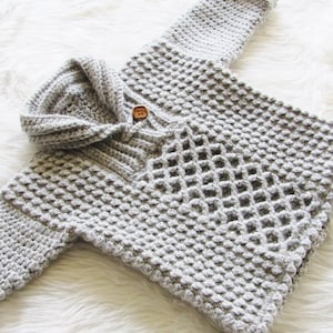 May include: A light gray, handmade crochet baby sweater with a hood and a single wooden button closure. The sweater features a textured stitch pattern and is displayed on a white, fluffy surface.