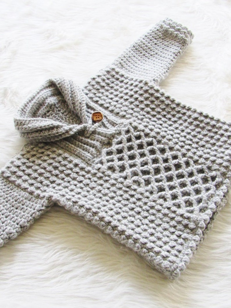 May include: A light gray, handmade crochet baby sweater with a hood and a single wooden button closure. The sweater features a textured stitch pattern and is displayed on a white, fluffy surface.