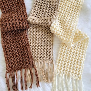 May include: Three rectangular crochet scarves in dark brown, light tan, and cream. Each scarf features an open, textured lattice stitch pattern and is finished with matching fringe tassels along the bottom edge of the fabric.
