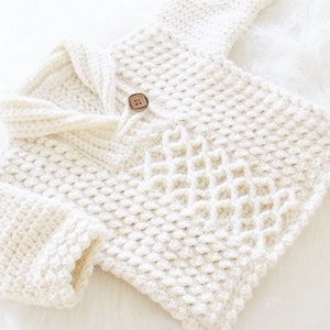 May include: Cream-colored, hand-knitted baby sweater with a textured diamond pattern on the front. The sweater features a shawl collar, long sleeves, and a wooden button closure. The soft, cozy knitwear is perfect for keeping a baby warm.