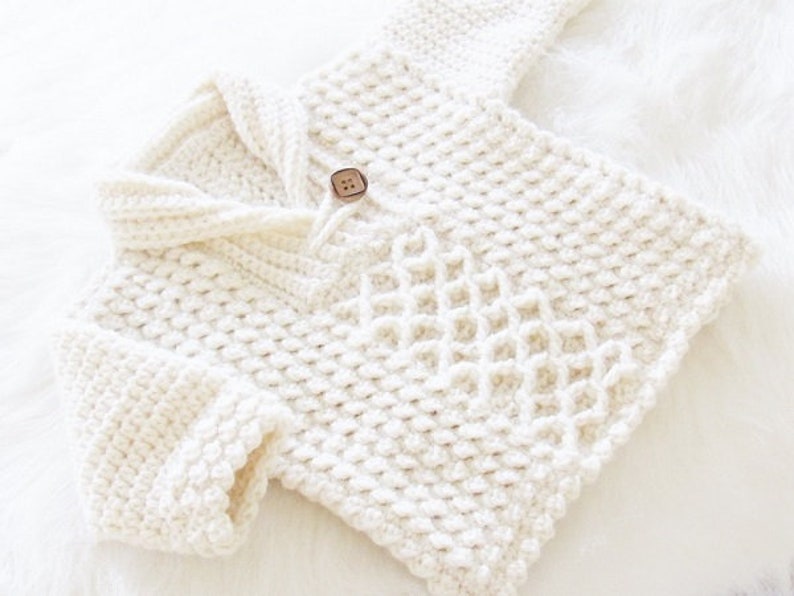 May include: Cream-colored, hand-knitted baby sweater with a textured diamond pattern on the front. The sweater features a shawl collar, long sleeves, and a wooden button closure. The soft, cozy knitwear is perfect for keeping a baby warm.