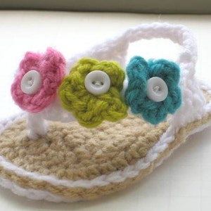 Könnte beinhalten: Gehäkelte Babysandalen mit einem weißen Riemen und drei Blumenverzierungen. Die Blumen sind rosa, grün und blau mit weißen Knöpfen in der Mitte.