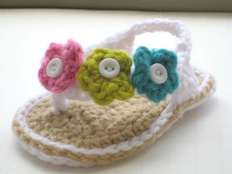 May include: Crocheted baby sandals with a white strap and three flower embellishments. The flowers are pink, green, and blue with white buttons in the center.