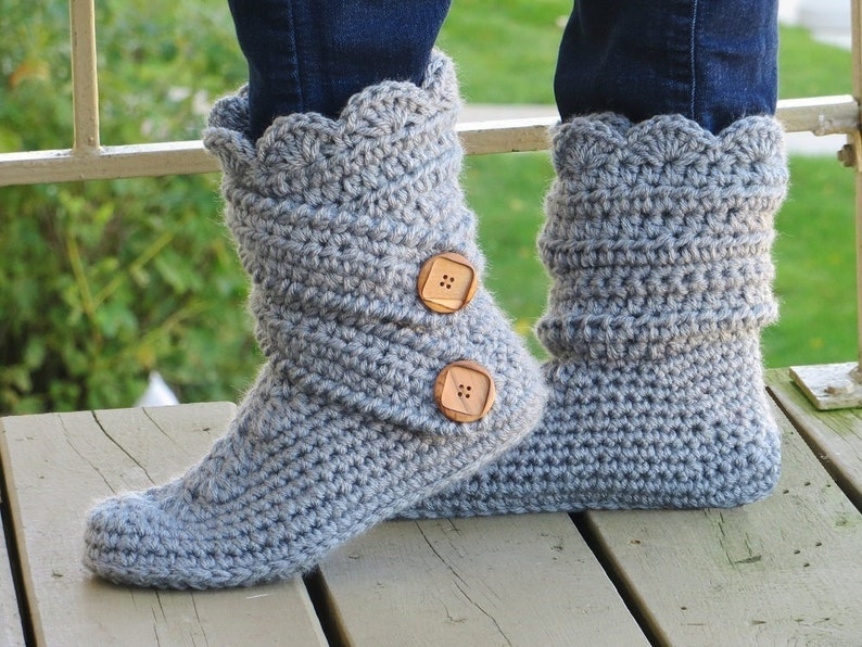 May include: A pair of gray crochet boots with a scalloped cuff and two wooden buttons. The handmade boots are worn over blue jeans. The boots appear to be soft and warm, perfect for indoor wear.