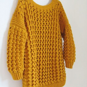 May include: A mustard yellow crocheted sweater with a loose fit and long sleeves. The sweater has a textured stitch pattern and a wide neckline.