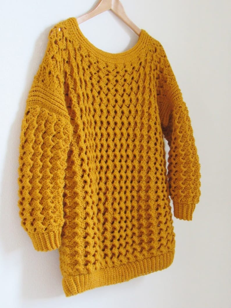 May include: A mustard yellow crocheted sweater with a loose fit and long sleeves. The sweater has a textured stitch pattern and a wide neckline.