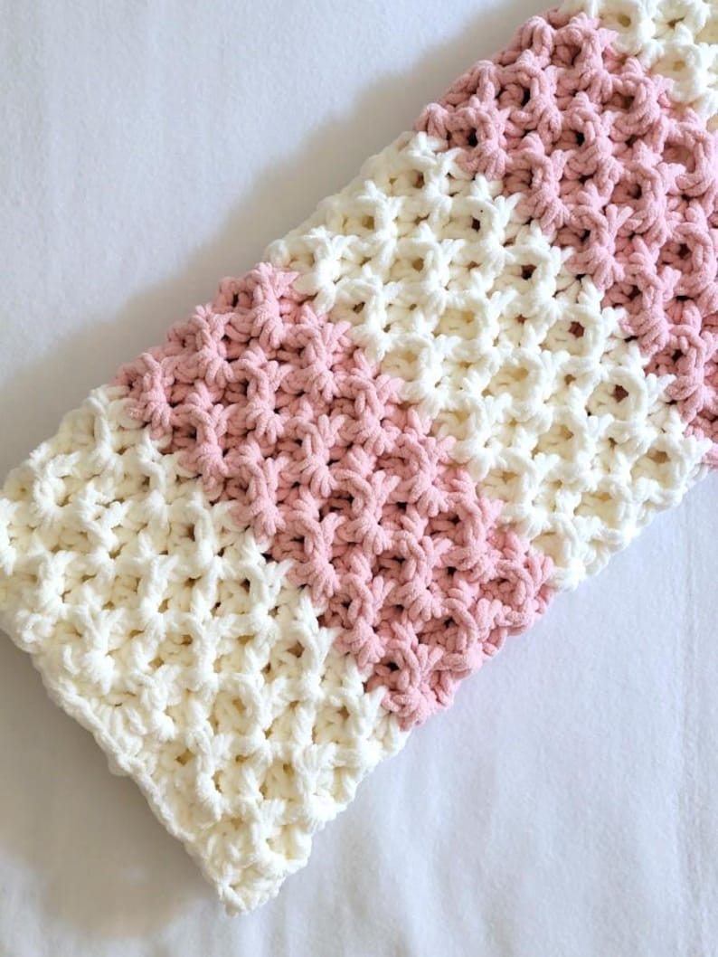 May include: A crocheted baby blanket with a white and pink striped pattern. The blanket is made with a soft, textured yarn.