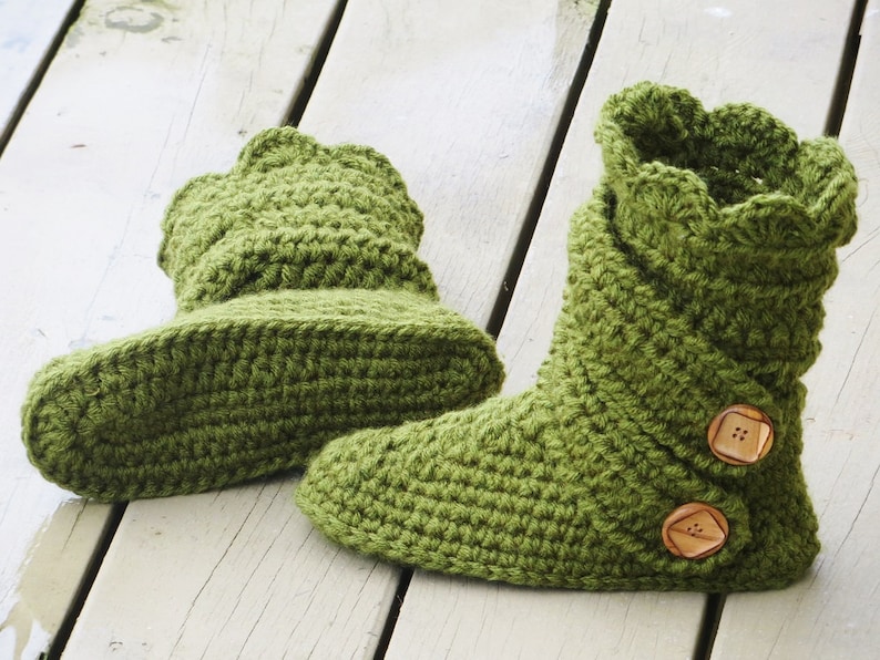 May include: A pair of handmade, green crochet boots. The boots have a scalloped top edge and are secured with two wooden buttons. The boots are made of a thick, textured yarn and are designed for indoor wear.