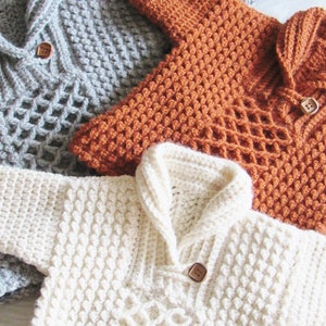 May include: Close-up of four handmade crochet sweaters in various colors. The sweaters are in shades of gray, orange, and white. Each sweater has a collar and a wooden button closure. The crochet work features textured patterns and openwork designs.