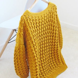 May include: A mustard yellow crocheted sweater with a loose fit and a wide neckline. The sweater is hanging on a wooden hanger.