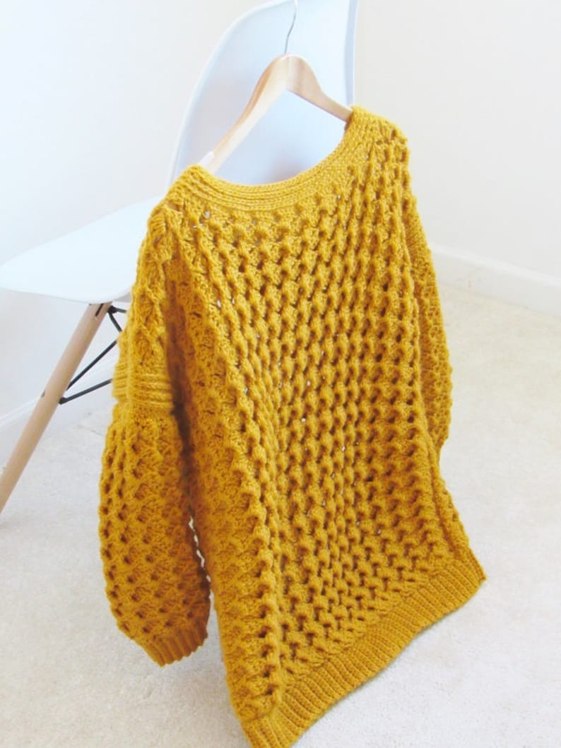 May include: A mustard yellow crocheted sweater with a loose fit and a wide neckline. The sweater is hanging on a wooden hanger.