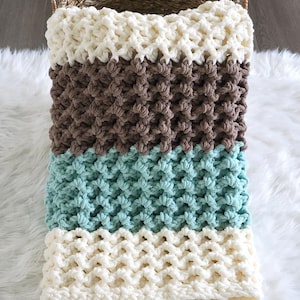 May include: A handmade, chunky knit blanket in a basket. The blanket features alternating bands of cream, brown, and teal colors. The texture is thick and the stitches are visible, creating a cozy, textured appearance. The blanket is resting on a white, fluffy surface.