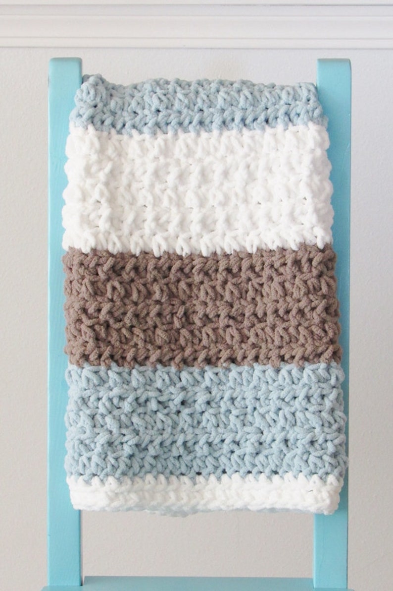 K&ouml;nnte beinhalten: Eine gestreifte geh&auml;kelte Babydecke in den Farben Blau, Braun und Wei&szlig;. Die Decke liegt &uuml;ber einem blauen Stuhl.