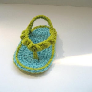 May include: A crocheted baby sandal in blue and green. The sandal has a strap that goes around the ankle and a toe post.