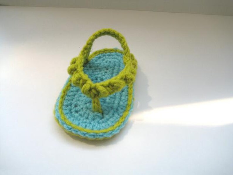 May include: A crocheted baby sandal in blue and green. The sandal has a strap that goes around the ankle and a toe post.