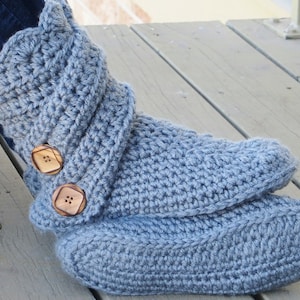 May include: A pair of light blue crochet slipper boots with two wooden buttons. The boots are handmade and appear soft and warm, perfect for indoor wear. The boots are resting on a wooden surface.