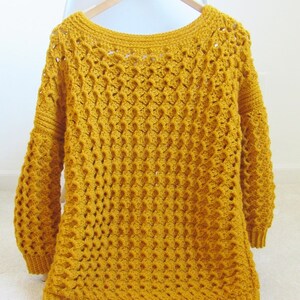 May include: A mustard yellow crocheted sweater with a loose fit and a wide neckline. The sweater has a textured stitch pattern and long sleeves.