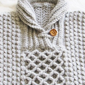 May include: A light gray, hand-crocheted sweater with a textured pattern. The sweater features a folded collar, a wooden button closure, and a decorative open-weave design on the front. The yarn appears soft and warm.
