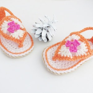 May include: A pair of crocheted sandals in orange and white with pink flower accents. The sandals have a strap that goes around the ankle.