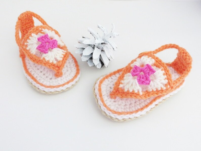 May include: A pair of crocheted sandals in orange and white with pink flower accents. The sandals have a strap that goes around the ankle.