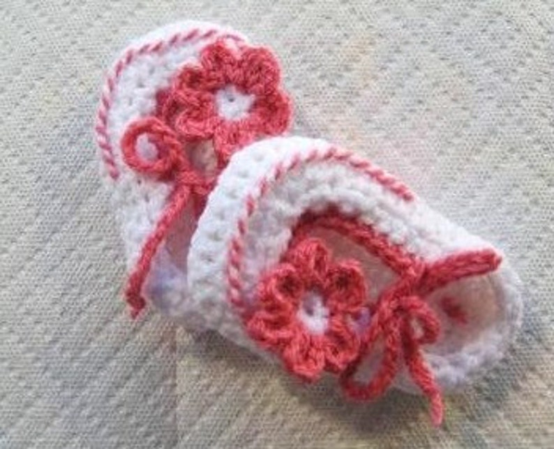 May include: A pair of white crocheted baby booties with pink flower accents and white ribbon ties.