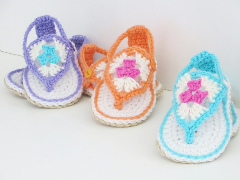 May include: Three pairs of crocheted baby sandals in purple, orange, and blue with white and pink flower accents.