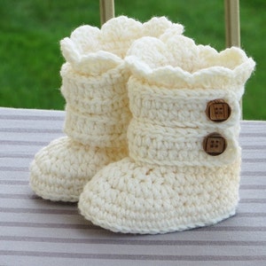 May include: A pair of white crocheted baby booties with a ruffled top and wooden button accents.