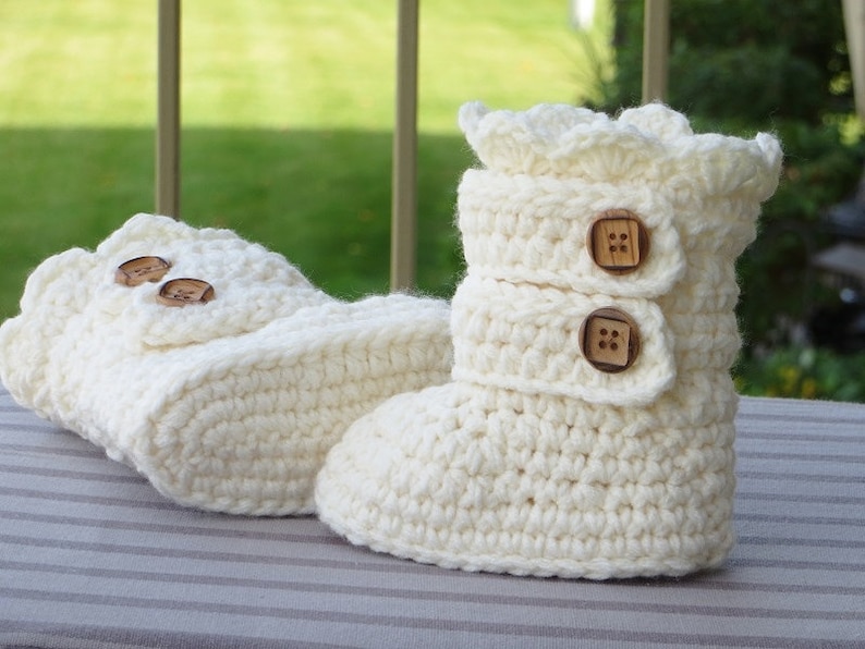 May include: A pair of white crocheted baby booties with wooden button closures. The booties have a ruffled top and are made with a soft, textured yarn.