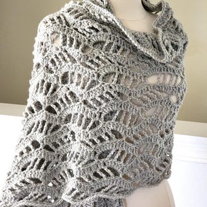 May include: A light grey, open-weave crochet shawl draped over a mannequin. The shawl features a repeating geometric pattern with a scalloped edge and a cowl neck. The handmade shawl is a soft, neutral accessory.