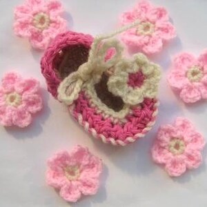 May include: A pink and cream crocheted baby shoe with a flower design. The shoe is surrounded by six smaller pink crocheted flowers.