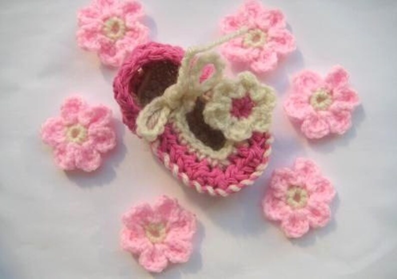 May include: A pink and cream crocheted baby shoe with a flower design. The shoe is surrounded by six smaller pink crocheted flowers.