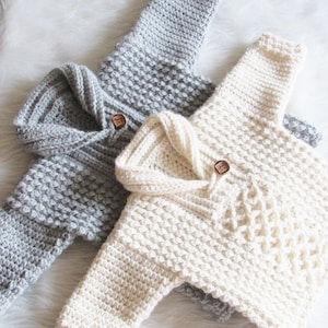 May include: Two handmade crochet baby sweaters. One is light gray, the other is off-white. Each has a hood, long sleeves, and a wooden button closure. The sweaters are made with a textured stitch.