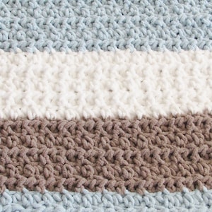 K&ouml;nnte beinhalten: Eine gestreifte Strickdecke aus braunem, hellblauem und wei&szlig;em Garn. Die Decke ist aus einem dicken Garn gefertigt und hat eine weiche, flauschige Textur.