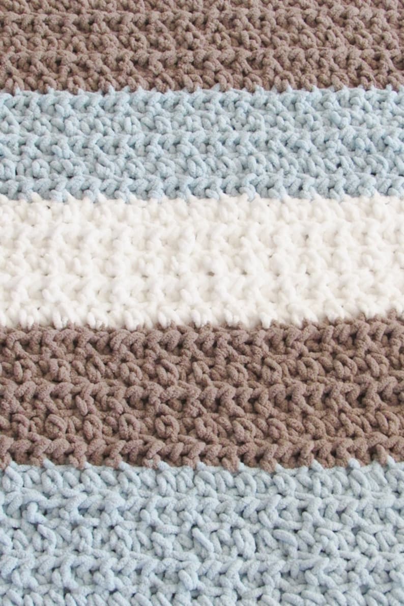 K&ouml;nnte beinhalten: Eine gestreifte Strickdecke aus braunem, hellblauem und wei&szlig;em Garn. Die Decke ist aus einem dicken Garn gefertigt und hat eine weiche, flauschige Textur.