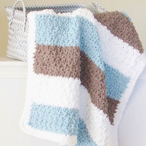 K&ouml;nnte beinhalten: Eine gestreifte geh&auml;kelte Babydecke in den Farben Blau, Braun und Wei&szlig;. Die Decke ist gefaltet und &uuml;ber einen geflochtenen Korb gelegt.