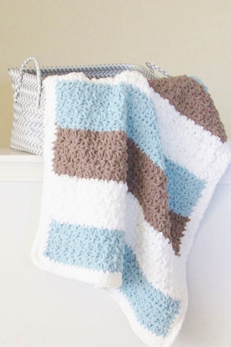 K&ouml;nnte beinhalten: Eine gestreifte geh&auml;kelte Babydecke in den Farben Blau, Braun und Wei&szlig;. Die Decke ist gefaltet und &uuml;ber einen geflochtenen Korb gelegt.
