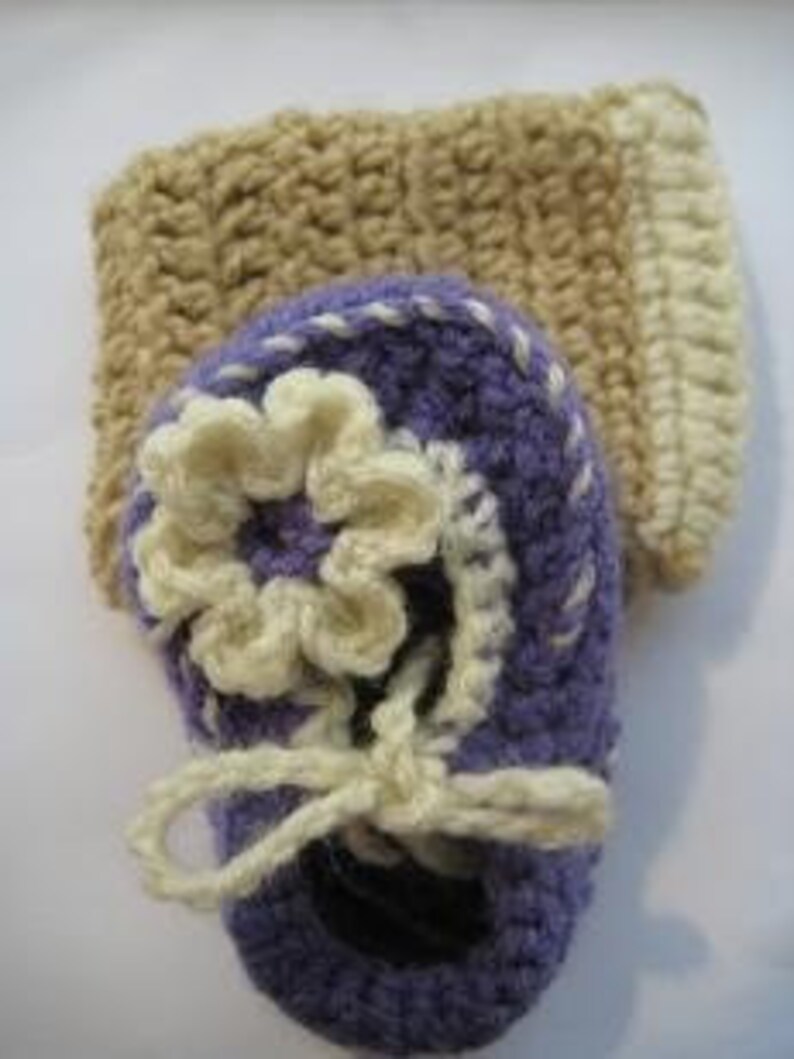 May include: Purple crocheted baby booties with a white flower and a beige crocheted leg warmer.