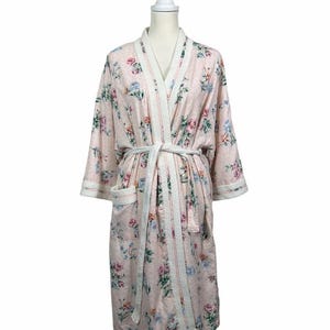 May include: A light pink robe featuring a floral pattern with roses in shades of pink, blue, and green. The garment includes a white quilted trim along the collar, cuffs, and pockets, and a matching white fabric tie belt at the waist.