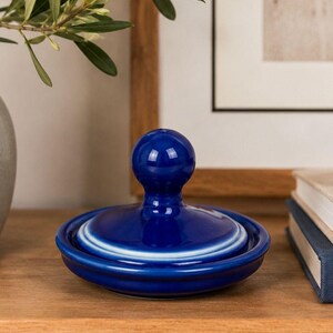 May include: A glossy cobalt blue ceramic lid featuring a smooth, rounded knob handle at the centre. The lid has a circular shape with a slightly raised rim and a subtle white gradient detail around the edge, showcasing a polished, reflective finish.